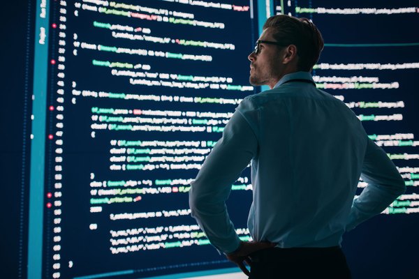 This image features a person standing with their back to the camera, looking at a large screen displaying lines of computer code. The person is wearing a light blue shirt and glasses, and appears to be focused on the content of the screen. The setting suggests a technology or data center environment, emphasizing a theme of software development or data analysis.