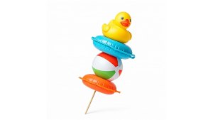 A creative composition featuring a skewer-like arrangement of beach-themed inflatable objects stacked vertically against a plain white background. At the top, a yellow rubber duck sits on a small blue inflatable ring. Below it, a classic multicolored beach ball with red, green, blue, and white segments is wedged between two additional inflatable rings, an orange one at the bottom and a blue one just beneath the duck. A wooden skewer pierces through all the objects, giving the appearance of a playful, oversized summer-themed kebab.